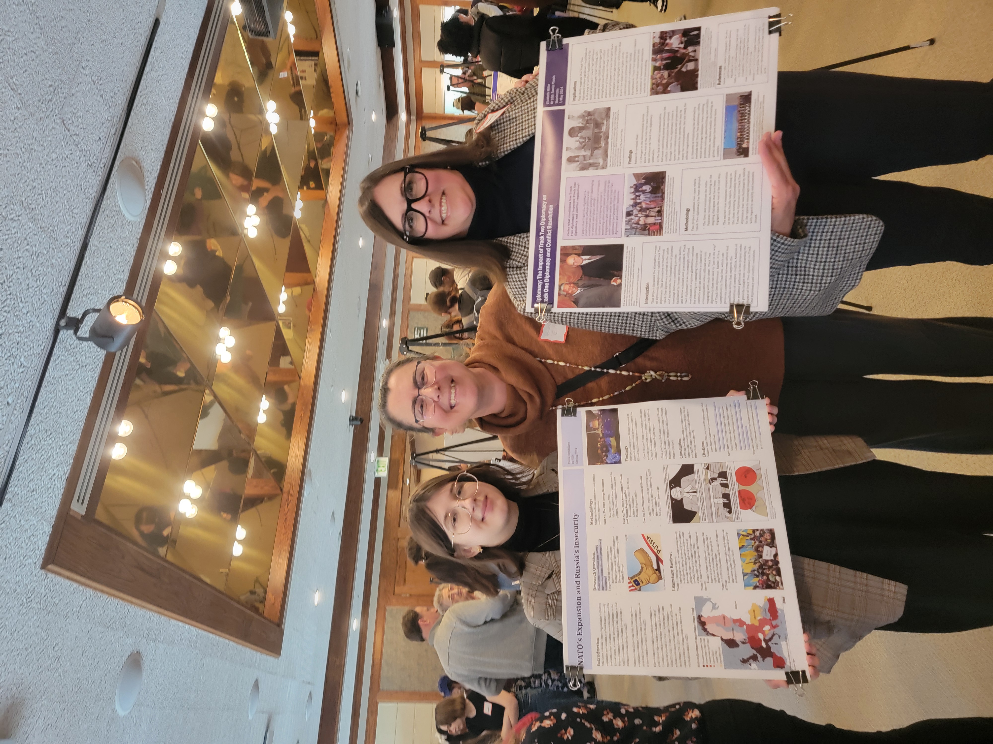 Two students in matching glasses, blazers, turtlenecks, and slacks stand and smile for the camera, holding their presentation posters. Professor Burcu Ellis stands between and behind them, smiling.