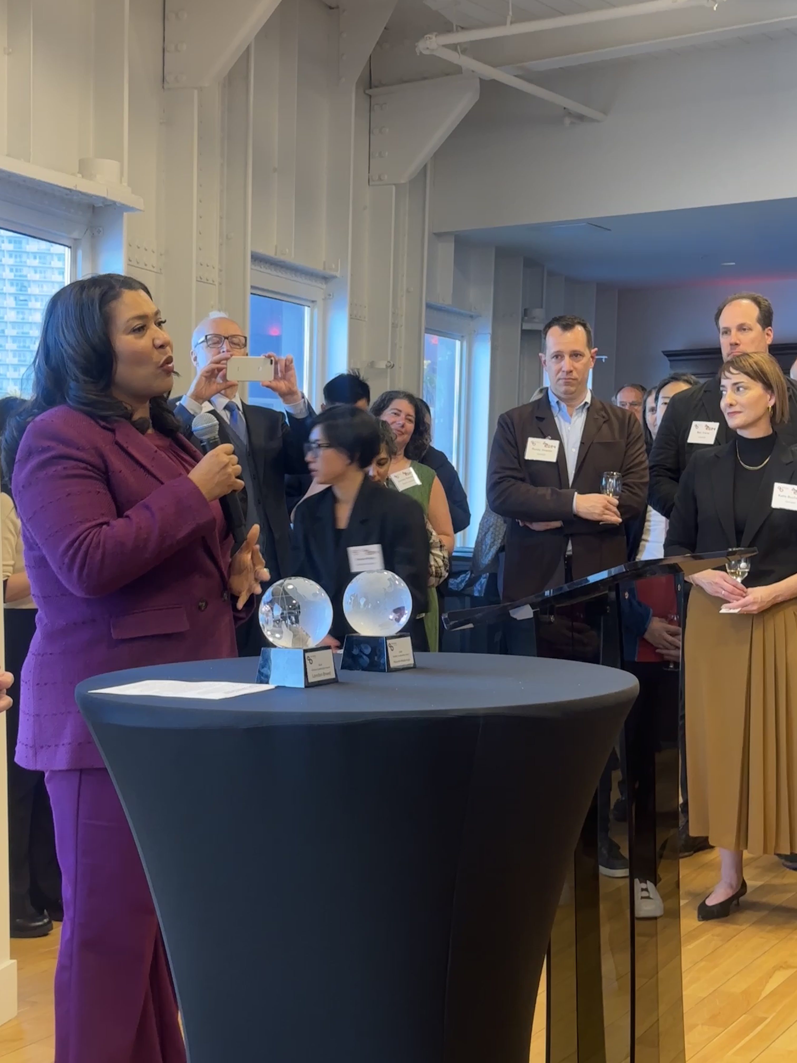SF Mayor London Breed holds a microphone and speaks to people in a conference room. She is presenting awards.