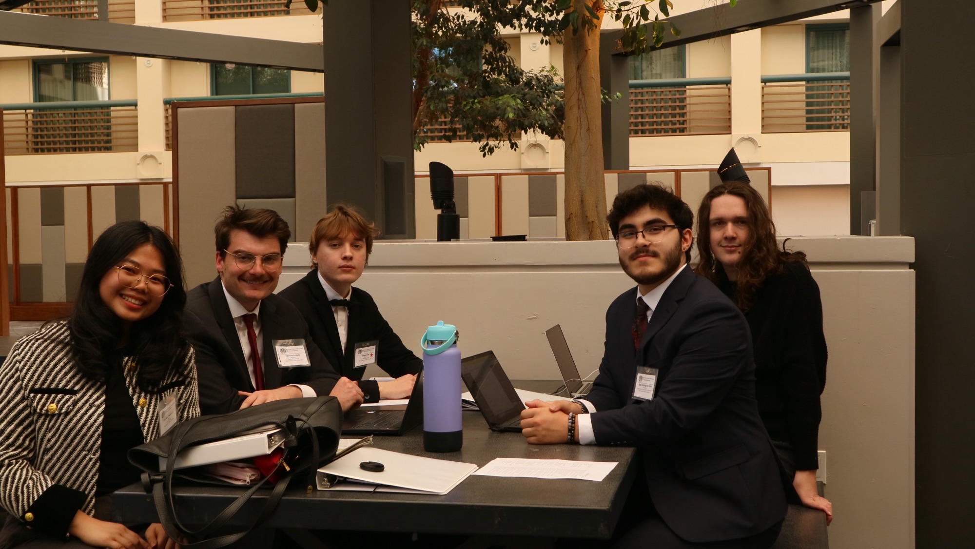 Five students sit at an outdoor table at the Model UN venue. They have laptops, water bottles, and papers ready. They are wearing suits. They face the camera and smile.