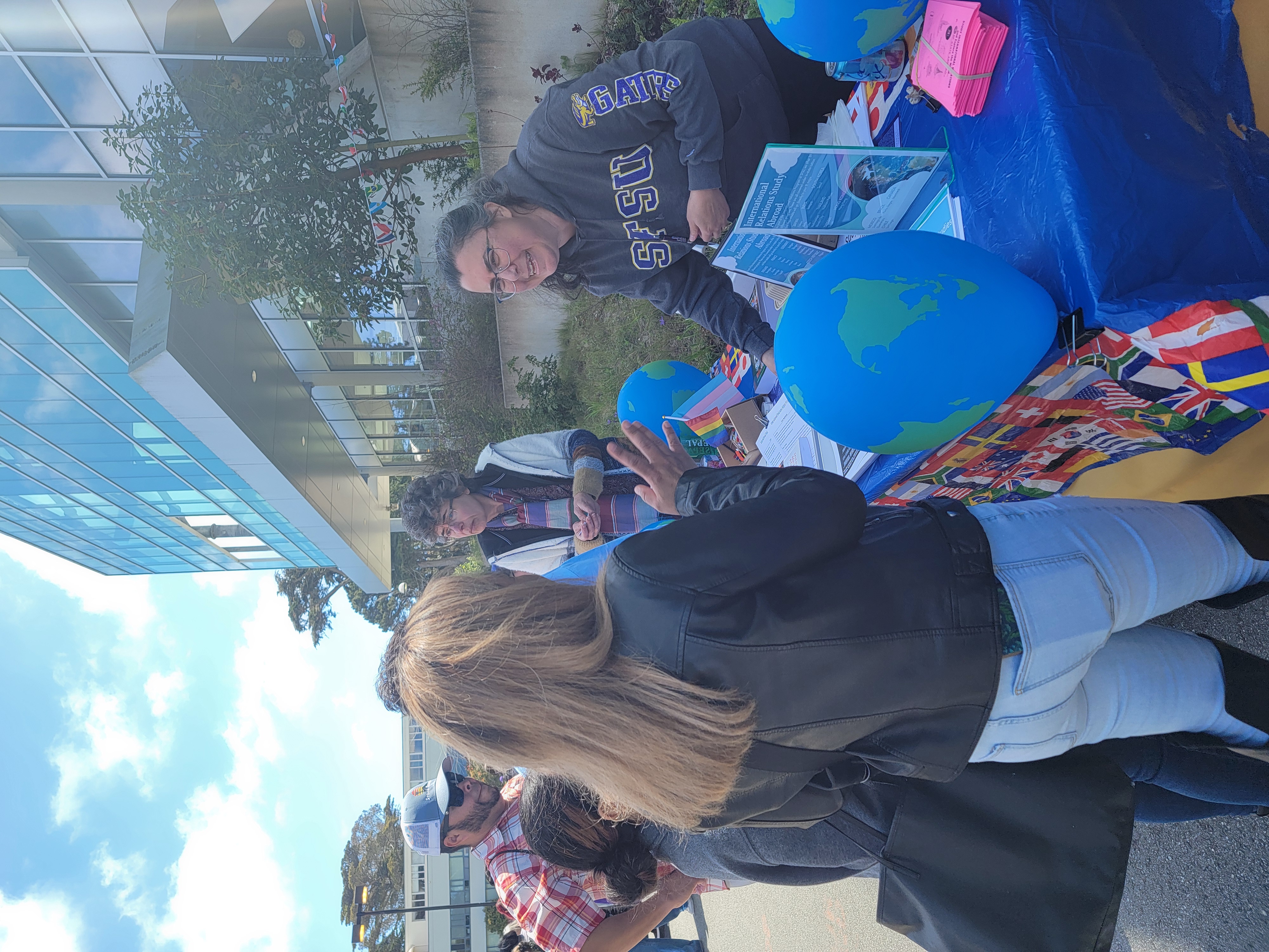 Professor Ellis speaks to a student at the International Relations Department table, which features globe-shaped balloons and flyers. Professor Volk stands nearby, speaking to another family.
