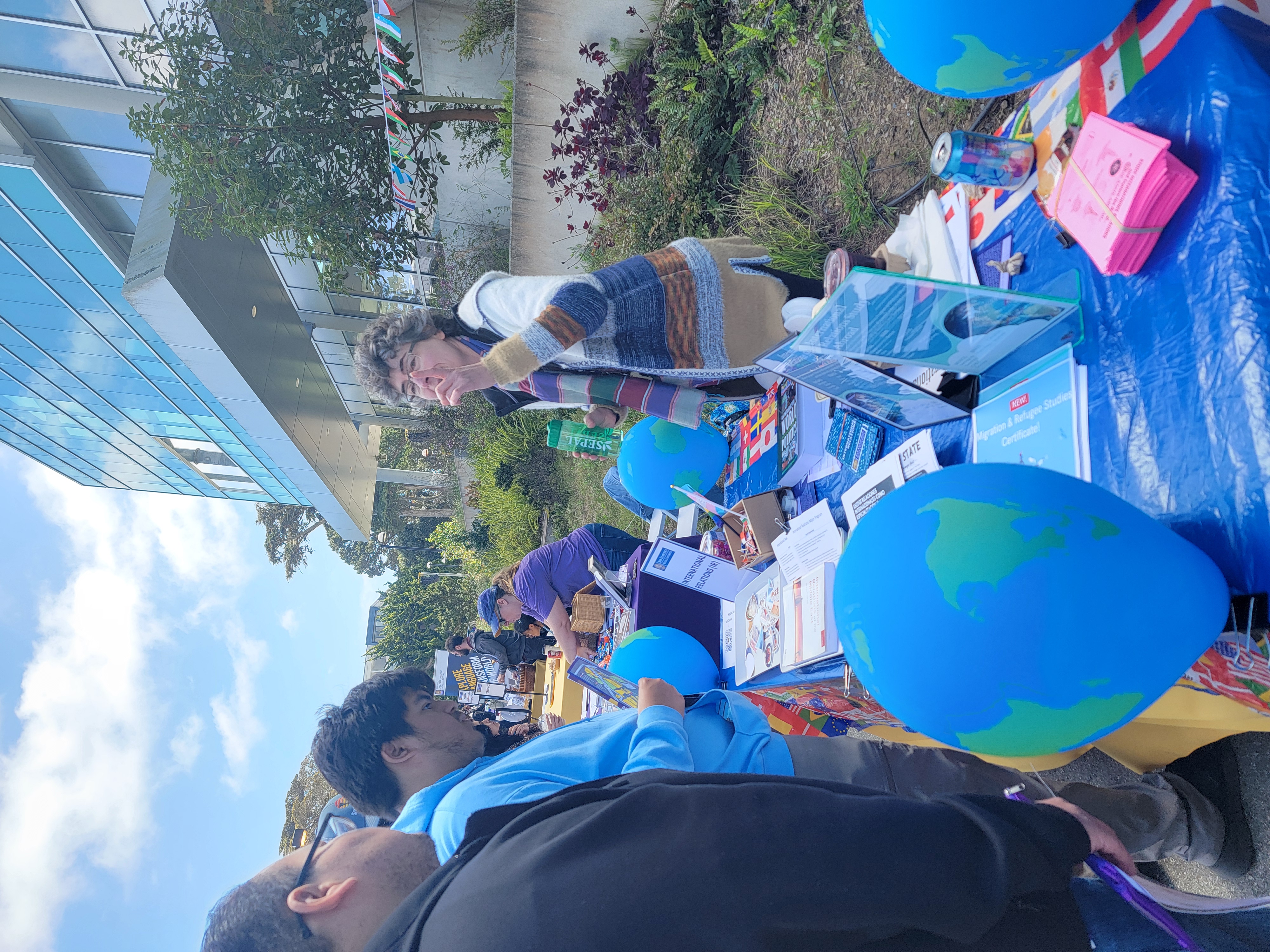Professor Volk stands behind the International Relations Department table, gesturing enthusiastically while speaking to a visiting student and parent. The table features globe-shaped balloons and flyers.