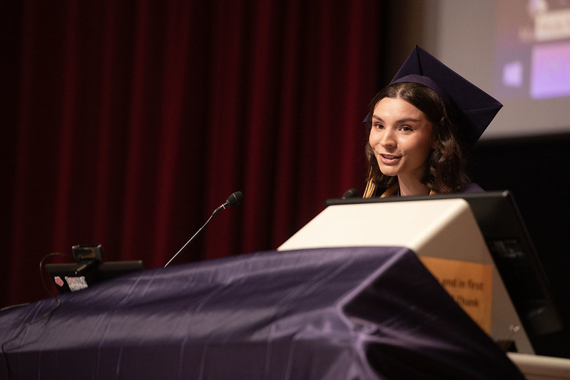 Faith Donithan gives a graduation speech. She looks to the left behind a podium. She is wearing a purple graduation cap and robe, as well as a yellow tassel and graduation stole.