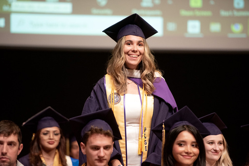 Zen Lewis stands above her peers, who are seated. She wears purple graduation regalia and a yellow stole over a white dress. She is smiling. Her peers, who are seated, also wear purple graduation regalia.